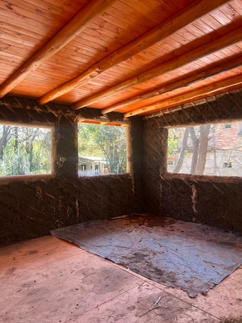 Interior de habitación en construcción. Paredes de material terroso rugoso, techo de madera con vigas de troncos. Tres ventanas abiertas. Piso de tierra rojiza con lona negra