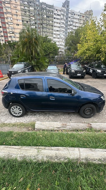 Vista lateral de un auto azul estacionado, con patrulleros de la Policía de la Ciudad y agentes al fondo, frente a edificios residenciales y árboles bajo cielo nublado