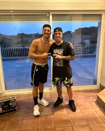 Dos hombres jóvenes sonríen; uno, sin camisa, señala al otro, que lleva gorra y camiseta negra con diseño. Ambos tienen shorts y zapatillas