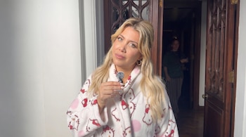 Mujer rubia con ojos claros, Wanda Nara, mirando a cámara, vestida con una bata blanca de Hello Kitty y sosteniendo un pequeño micrófono gris. De fondo, una puerta de madera.