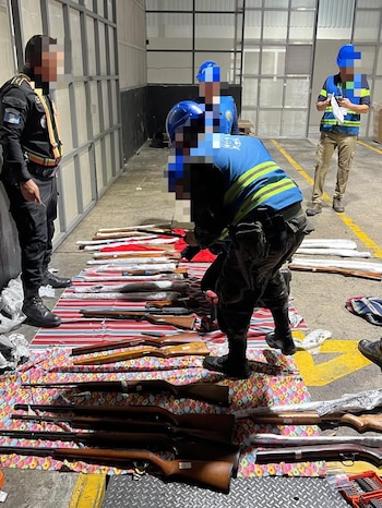 Interior de un almacén con varias personas en cascos azules y chalecos reflectantes. Inspeccionan y organizan cajas de cartón apiladas en palets