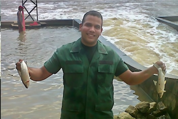 Hombre de tez morena y cabeza rapada, vestido con camisa verde, sostiene un pez en cada mano. Fondo de río con corrientes y otra persona en el agua