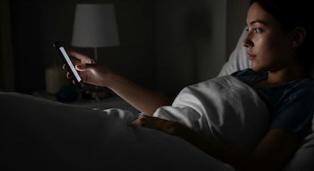 Mujer joven acostada en la cama en una habitación oscura, con la pantalla brillante de su teléfono móvil iluminando su rostro y mano.
