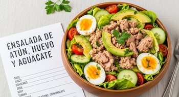 Un tazón de madera lleno de ensalada de atún, aguacate en rodajas, huevos cocidos por la mitad, lechuga, tomates cherry y rodajas de pepino, junto a una tarjeta de receta.