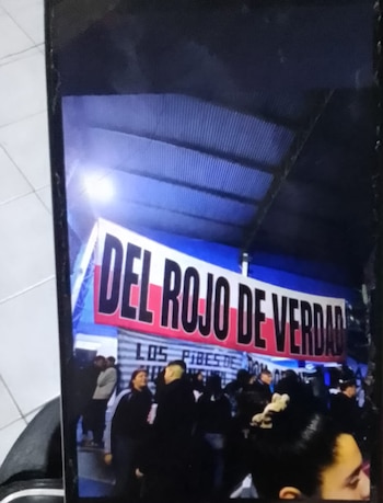 Una bandera con los colores rojo y blanco y la frase 'DEL ROJO DE VERDAD' cuelga bajo un techo metálico azul, con personas reunidas debajo