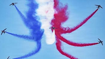 Siete aeronaves dejando estelas de humo azul, blanco y rojo en el cielo claro. La estela blanca emerge del avión central
