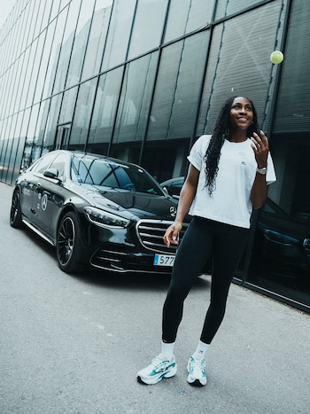 Una mujer con cabello trenzado, camiseta blanca y leggings negros sonríe mientras lanza una pelota de tenis. Detrás, un sedán Mercedes-Benz negro y un edificio de vidrio
