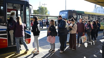 Vista de un grupo de personas esperando y abordando un autobús azul y amarillo en una parada, bajo la luz del sol