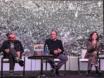 Tres personas sentadas en un escenario frente a una pantalla grande. Una mujer a la derecha sostiene un micrófono. Un libro y una botella de agua sobre una mesa redonda
