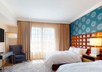 Una habitación de hotel luminosa con dos camas, cabecero de piel marrón, pared azul estampada, ventanales con cortinas beige, un sillón oscuro y un escritorio