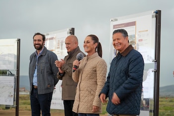 Claudia Sheinbaum, con micrófono, sonríe entre tres hombres, todos en chaquetas, de pie frente a paneles informativos bajo un cielo nublado