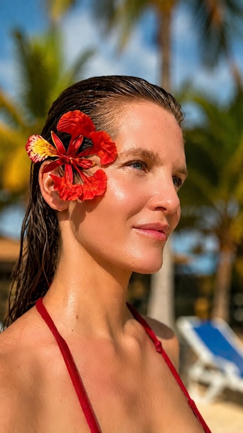 Liz Solari de perfil con el cabello mojado, una flor roja sobre la oreja y palmeras desenfocadas de fondo