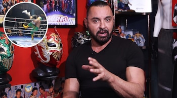 Un hombre con camiseta negra habla frente a tres máscaras tradicionales y un círculo insertado con una escena de lucha libre.