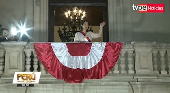 Una mujer con banda bicolor saluda desde un balcón adornado con la bandera peruana, junto a personas con cámaras fotográficas y destellos de flashes