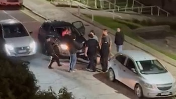 Vista aérea nocturna de un grupo de personas de pie alrededor de un SUV oscuro con la puerta abierta en una calle, con un coche blanco estacionado cerca