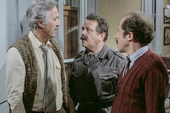 Tres hombres de mediana edad a mayores, uno con chaleco de punto, otro con camisa militar y un tercero con cárdigan, conversan en un interior