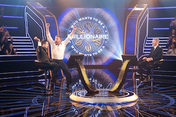 Estudio de televisión con tres hombres. El central eleva los brazos, los otros dos sonríen. Pantalla con el logo 'Who Wants to Be a Millionaire'. Público y luces