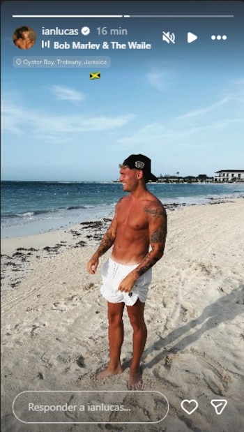 Un hombre sin camiseta, con bañador blanco y gorra negra, de pie en una playa de arena clara. Al fondo hay un mar azul con olas suaves y edificaciones