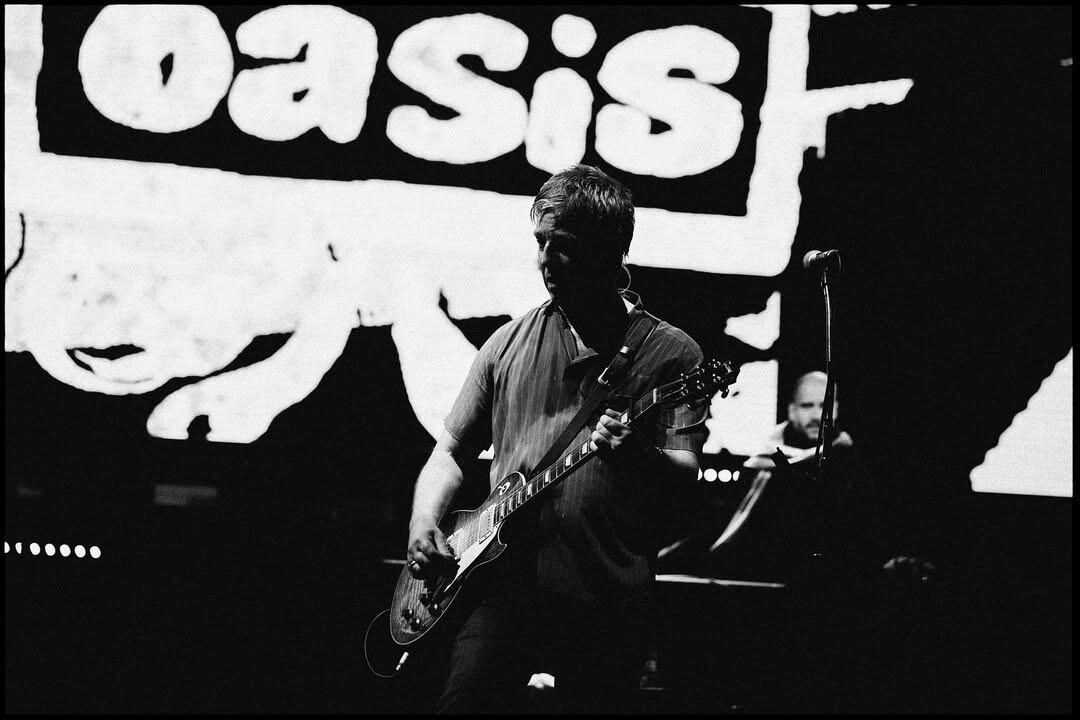 Noel Gallagher toca su guitarra bajo una luz tenue que resalta su figura en pleno escenario