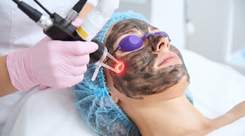 Mujer acostada con mascarilla negra y gafas moradas mientras un profesional aplica un dispositivo de láser con luz roja en su mejilla.