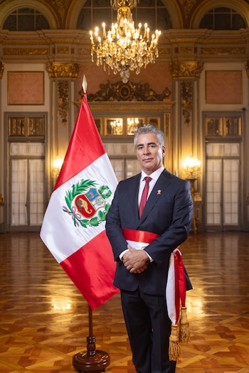 Un hombre de traje oscuro, corbata roja y banda bicolor posa de pie junto a una bandera de Perú en un salón con piso de madera y candelabros