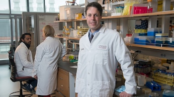 Un hombre con bata de laboratorio blanca y el logo de UMass Amherst sonríe a la cámara en un laboratorio. Dos colegas y equipo científico al fondo.