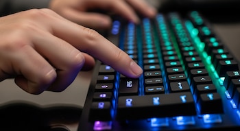 Primer plano de una mano sobre un teclado gaming negro retroiluminado con luces LED azules y verdes. Un dedo presiona la tecla "Shift".