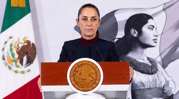 Mujer con chaqueta oscura y cuello burdeos. Detrás, un atril con el escudo de Estados Unidos Mexicanos, la bandera de México y una ilustración femenina.