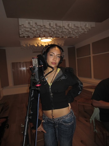 Una mujer joven con cabello oscuro recogido, usando auriculares y una sudadera negra sobre un top amarillo, posa de pie frente a un micrófono en un estudio de grabación