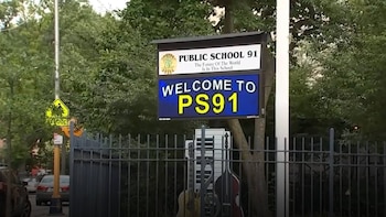 Cartel azul digital 'WELCOME TO PS91' y cartel de 'Public School 91' sobre una valla con instrumentos musicales. Árboles verdes y calle con coches a la izquierda