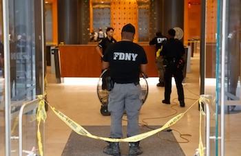 Un hombre de espaldas con camiseta con las siglas FDNY y un policía se encuentran tras una cinta amarilla de seguridad en la entrada de un edificio