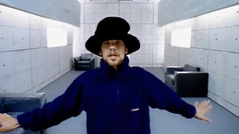 Jamiroquai, con sombrero oscuro y ropa azul y negra, caminando en una habitación gris con paredes de concreto y sofás negros