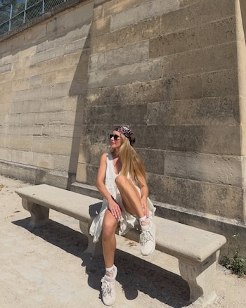 Una mujer con cabello rubio sentada en un banco de piedra, viste un vestido blanco, un pañuelo estampado y gafas de sol, frente a una pared de bloques