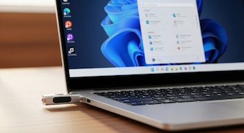 Una memoria USB plateada insertada en el puerto lateral de una laptop. La pantalla muestra el escritorio de Windows 11 con el menú de inicio abierto.