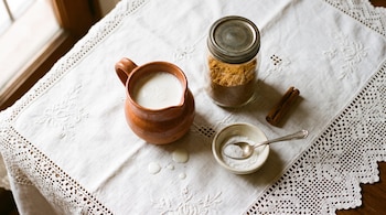 Jarra de barro con leche, frasco de vidrio con azúcar, canela y un tazón con polvo blanco sobre un mantel de lino bordado.
