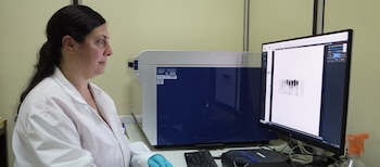 Una mujer con bata de laboratorio y guantes azules observa una pantalla de computadora junto a un equipo de análisis