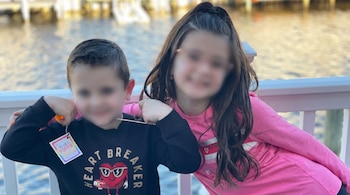 Dos niños con caras borrosas, un chico con sudadera negra y una chica con camiseta rosa, posan junto a una barandilla blanca con agua de fondo.