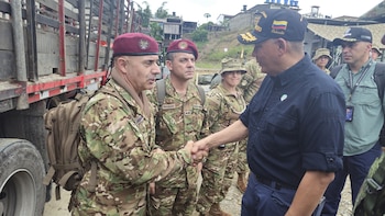 Un hombre con gorra de Colombia estrecha la mano de un militar con boina granate y uniforme de camuflaje junto a un camión
