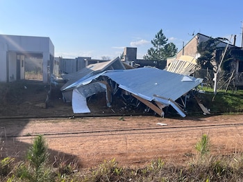 Techo de chapa y estructura de madera colapsados sobre un terreno de tierra. Se observan edificios distantes y cielo azul