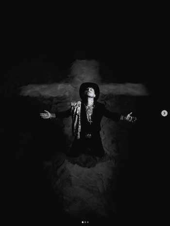 Forajido arrodillado en blanco y negro, brazos extendidos, cabeza hacia arriba, sombrero y joyas. Una cruz de luz se forma detrás de él en un fondo oscuro