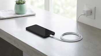 Fotografía editorial de un teléfono móvil negro conectado a un cargador blanco sobre una superficie gris de concreto, con el cable enrollado y un enchufe en la pared.