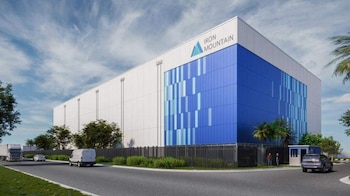 Edificio blanco y azul de Iron Mountain con un camión, una furgoneta y un coche en la carretera frontal, bajo un cielo parcialmente nublado