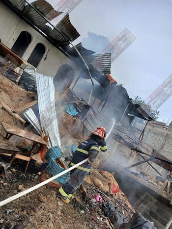 Un bombero con casco y uniforme negro y amarillo rocía agua con una manguera blanca sobre los escombros de una casa dañada