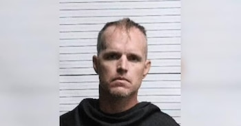 Ficha policial de un hombre de piel clara, cabello corto y prenda negra, posando de frente ante una escala de estatura