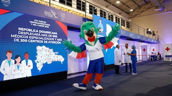 La mascota oficial de los Juegos Santo Domingo 2026, un perico, posa sonriente frente a una gran pantalla LED con un mapa de la República Dominicana y gráficos médicos.