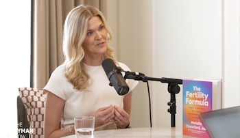 Mujer rubia sentada con micrófono, vaso de agua y un libro titulado "The Fertility Formula" en una mesa