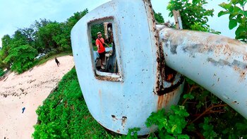 Hombre sonriente dentro de una estructura de cañón naval oxidado, ubicado en una playa arenosa con vegetación y árboles al fondo