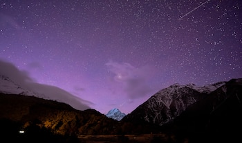 Cielo nocturno con estrellas y tonos púrpuras sobre montañas con cimas nevadas y una colina boscosa en primer plano
