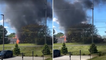Gran columna de humo negro y llamas anaranjadas se eleva sobre un área arbolada con cielo azul. Un coche y parte de una calle son visibles abajo
