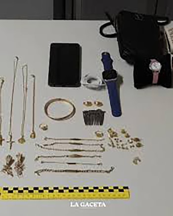 Varias cadenas de oro, pulseras, anillos, un teléfono móvil, dos relojes y una regla métrica amarilla y negra sobre una mesa blanca